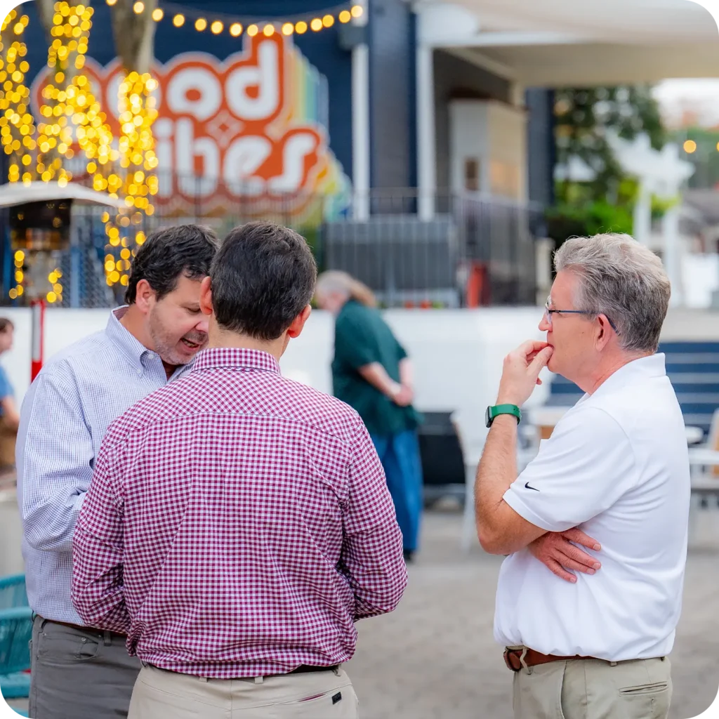 Three men talking together at an outdoor social event with decorative lights in the background.