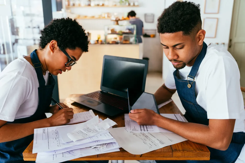 Two café owners reviewing paperwork and receipts together at a table with a laptop.