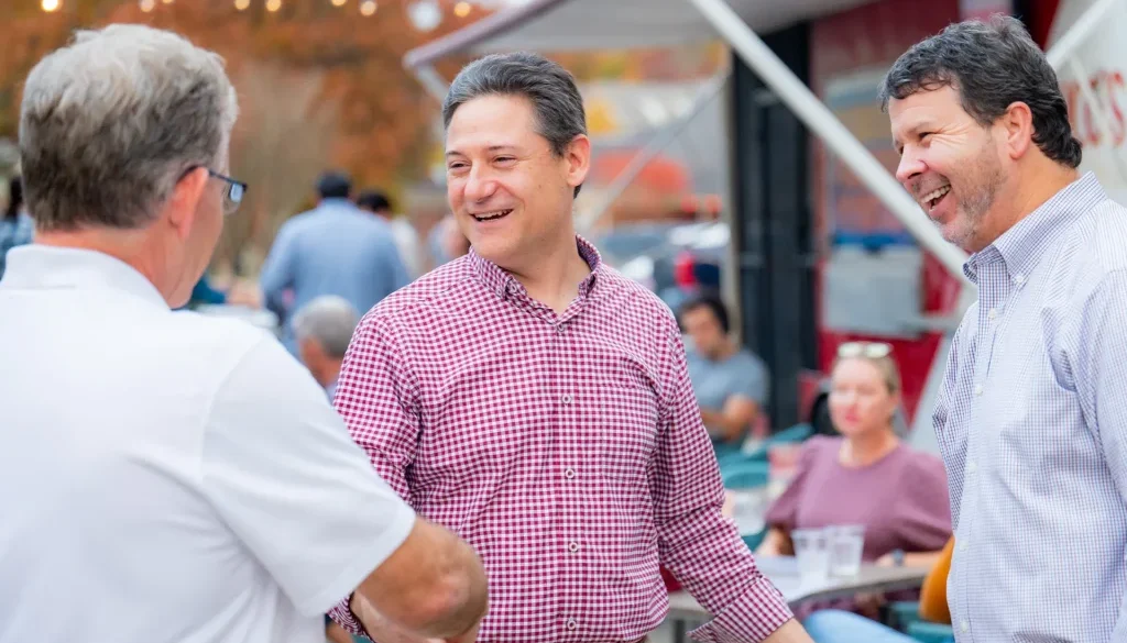Three men chatting and smiling at an outdoor anniversary event, with string lights and other guests in the background.