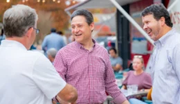 Three men chatting and smiling at an outdoor anniversary event, with string lights and other guests in the background.