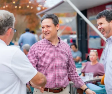Three men chatting and smiling at an outdoor anniversary event, with string lights and other guests in the background.
