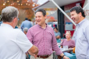 Three men chatting and smiling at an outdoor anniversary event, with string lights and other guests in the background.