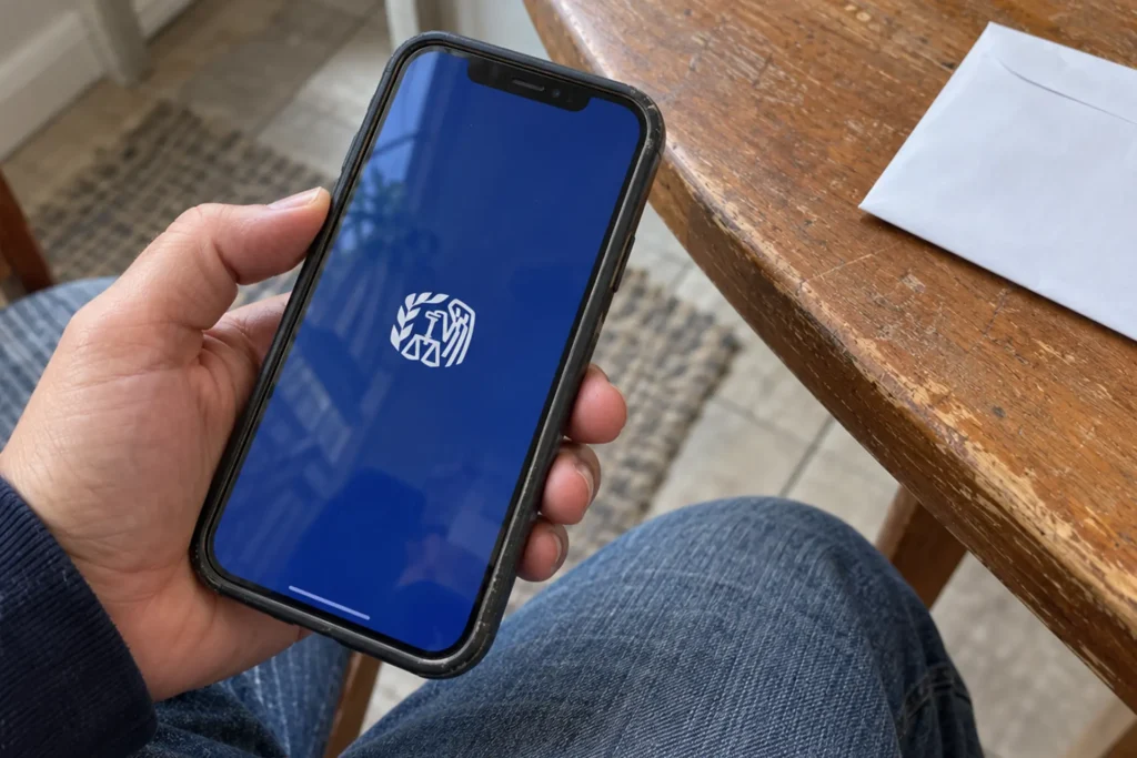Person holding a smartphone displaying the IRS emblem while seated at a kitchen table.